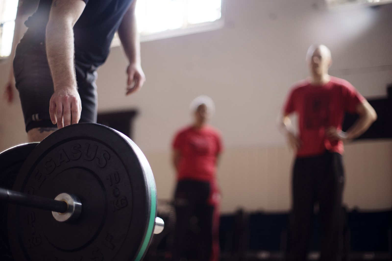 A loaded barbell in the foreground, two people standing behind it in a training room.